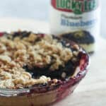 A blueberry pie in a glass pie plate missing a piece of pie. A can of Lucky Leaf Blueberry Pie Filling in the background.