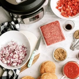 An Instant Pot, chopped red peppers, diced onions, ground beef, sauces, brown sugar, and burger buns are arranged on a countertop for meal preparation.