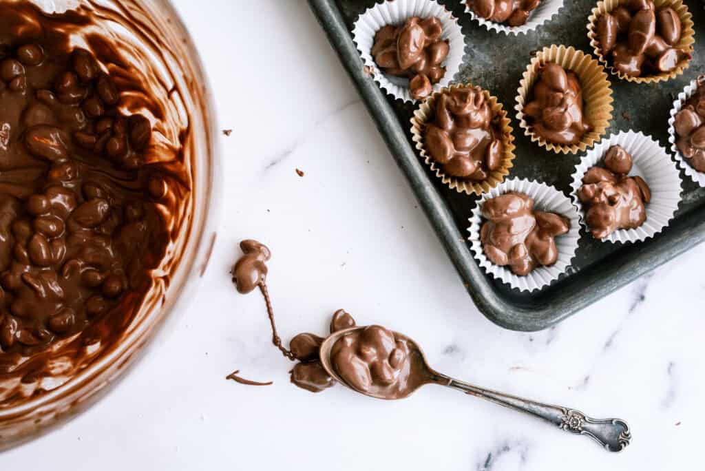 A spoon with chocolate on the counter. A sheet pan filled with cupcake liners that have chocolate nut clusters in them. An instant pot with a glass bowl of melted chocolate inside.