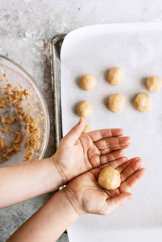 Peanut butter dough rolled into balls and placed on a baking sheet lined with wax paper. Two hands holding one peanut butter ball.