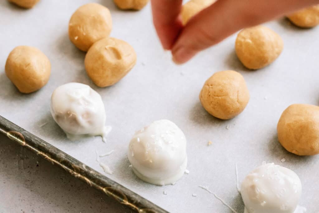 A baking sheet lined with wax paper with peanut butter balls and some Peanut Butter Snowballs with a hand sprinkling sprinkles onto a chocolate covered ball.