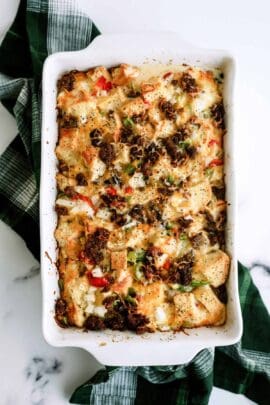 Baked casserole with bread cubes, bell peppers, onions, melted cheese, and browned sausage in a white rectangular dish on a green checkered cloth.