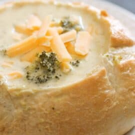 A bread bowl filled with creamy broccoli and cheese soup, garnished with shredded cheese.