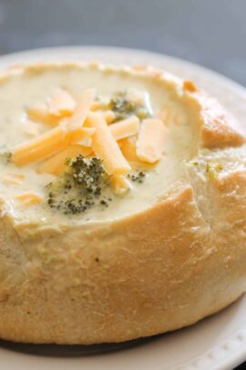 A bread bowl filled with creamy broccoli and cheese soup, garnished with shredded cheese.