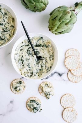 A bowl of spinach artichoke dip with a spoon, surrounded by crackers and two whole artichokes on a marble surface.