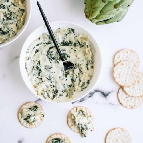 A bowl of spinach artichoke dip with a spoon, surrounded by crackers and two whole artichokes on a marble surface.