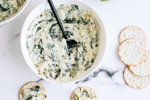 A bowl of spinach artichoke dip with a spoon, surrounded by crackers and two whole artichokes on a marble surface.