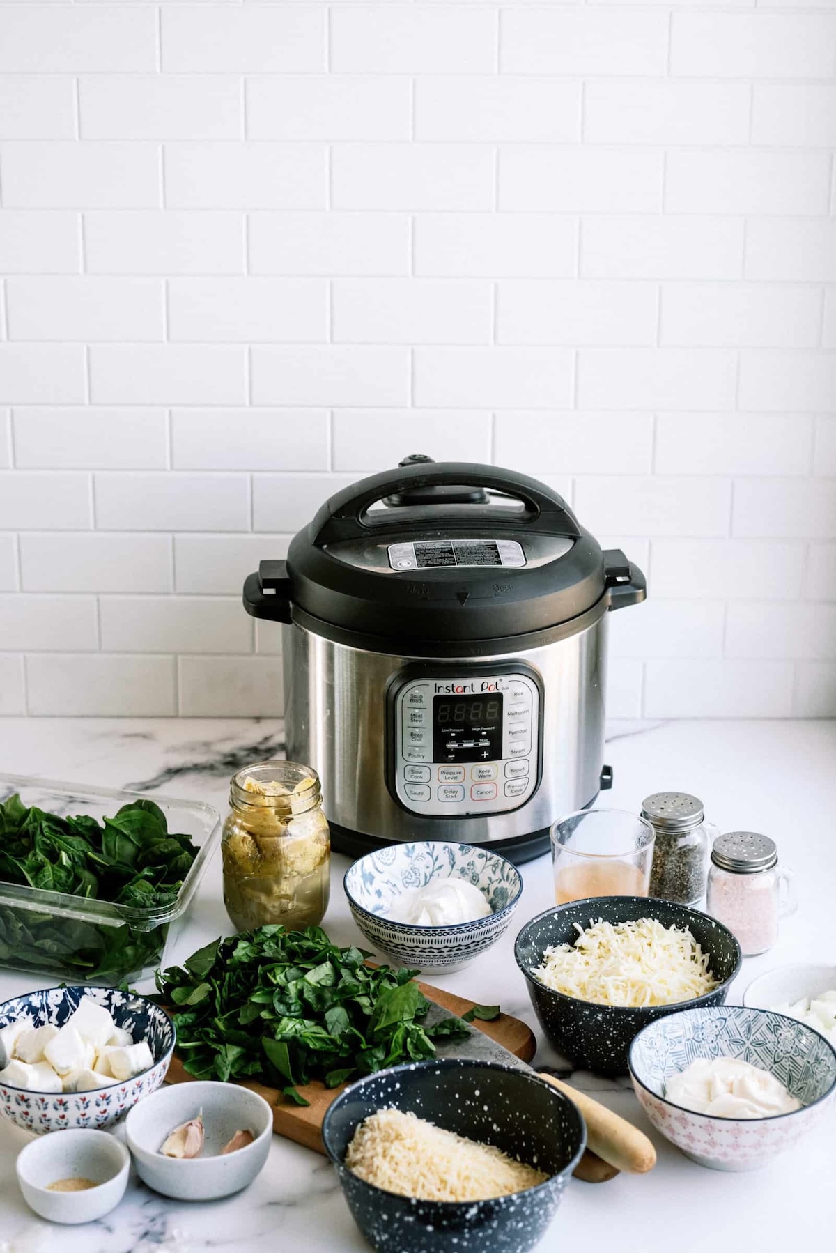 An electric pressure cooker on a counter surrounded by ingredients in bowls, including greens, cheese, spices, and a jar of artichokes.