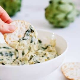 A hand holding a cracker dipped in a bowl of creamy spinach artichoke dip, with artichokes in the background.