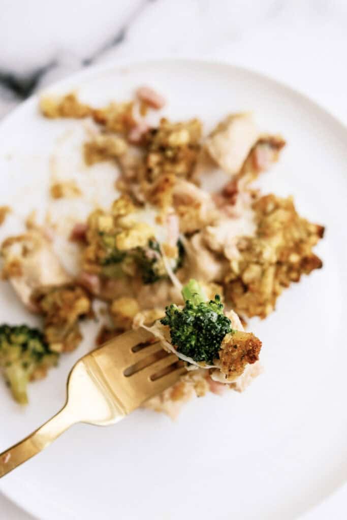 A fork holding a bite of a broccoli and chicken casserole with cheese, with additional portions of the casserole visible on a white plate.