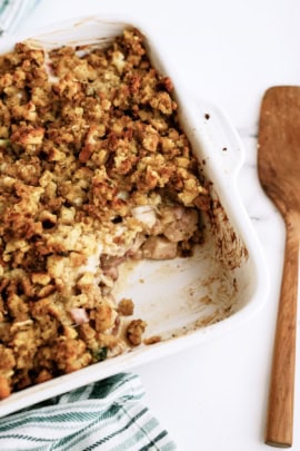 A white baking dish containing a partially eaten casserole with a breadcrumb topping, positioned next to a wooden spatula and a striped cloth on a marble surface.