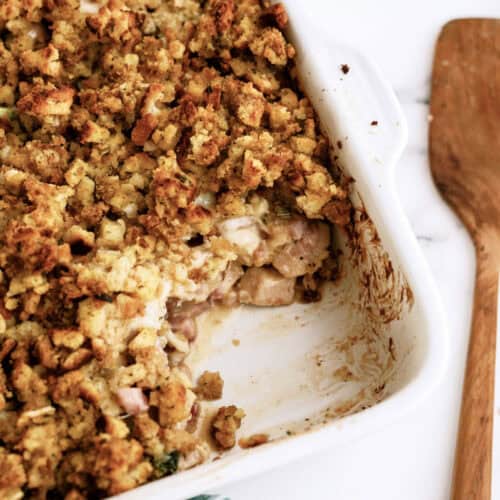 A white baking dish containing a partially eaten casserole with a breadcrumb topping, positioned next to a wooden spatula and a striped cloth on a marble surface.