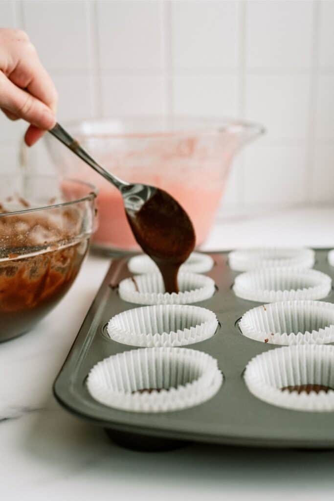 Spooning brownie batter into cupcake liners in cupcake tin. With a bowl of brownie batter off to the side.