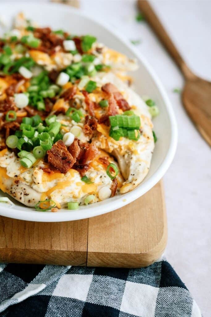 A white bowl filled with a baked potato dish topped with chopped green onions, bacon, and melted cheese sits on a wooden cutting board next to a blue and white checkered cloth.