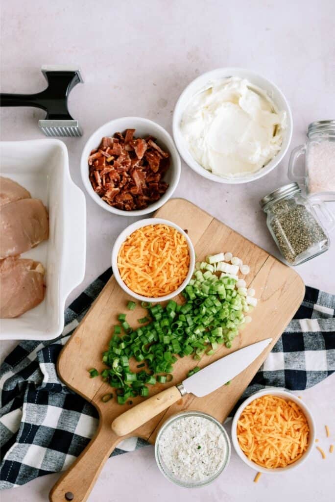 A kitchen countertop with ingredients for a recipe, including diced green onions, shredded cheese, chopped bacon, seasoning, raw chicken breasts, cream cheese, and a meat tenderizer.