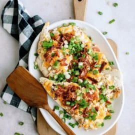 A plate of loaded baked potato slices topped with cheese, bacon, and green onions, placed on a wooden cutting board next to a wooden spatula and a black and white checkered cloth.