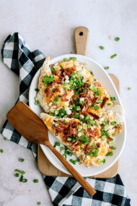 A plate of loaded baked potato slices topped with cheese, bacon, and green onions, placed on a wooden cutting board next to a wooden spatula and a black and white checkered cloth.