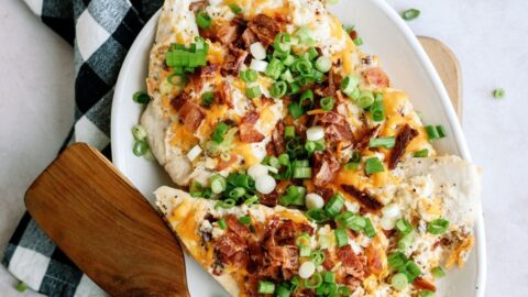 A plate of loaded baked potato slices topped with cheese, bacon, and green onions, placed on a wooden cutting board next to a wooden spatula and a black and white checkered cloth.