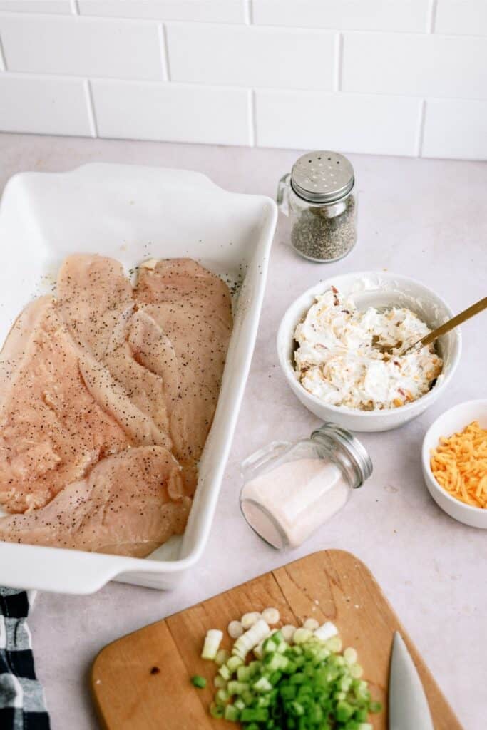 Raw seasoned chicken breasts in a white dish, a bowl with a creamy mixture and spoon, a small bowl of shredded cheese, chopped green onions on a cutting board with a knife, and a pepper shaker.