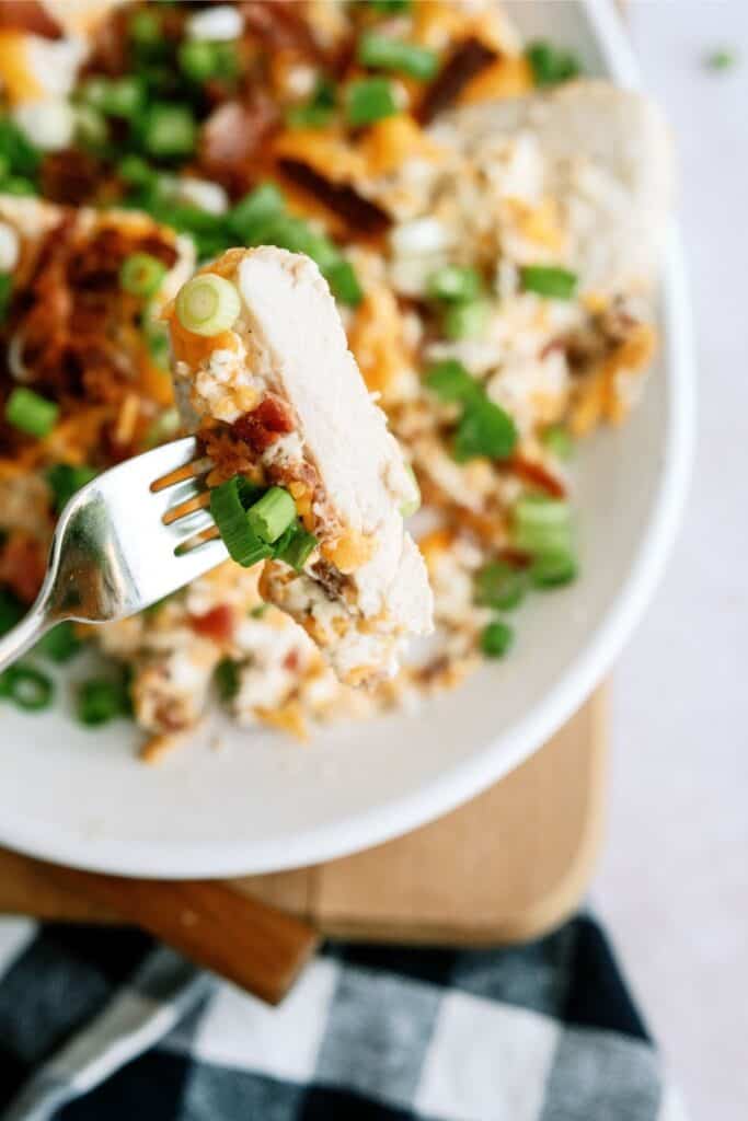 A fork holds a piece of food topped with chopped green onions and cheese over a dish of similar food, placed on a wooden surface next to a black and white checkered cloth.