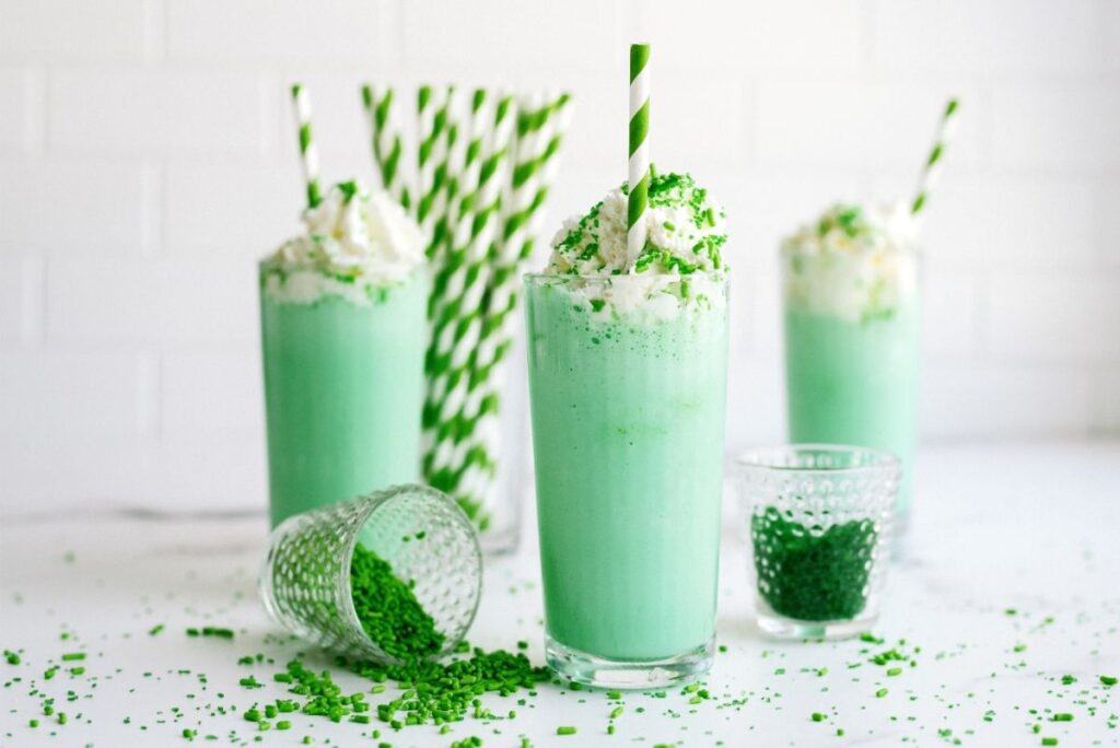 3 glasses with Copycat McDonald’s Shamrock Shakes in them. A small cup of sprinkles tipped over and a container holding striped green and white straws.