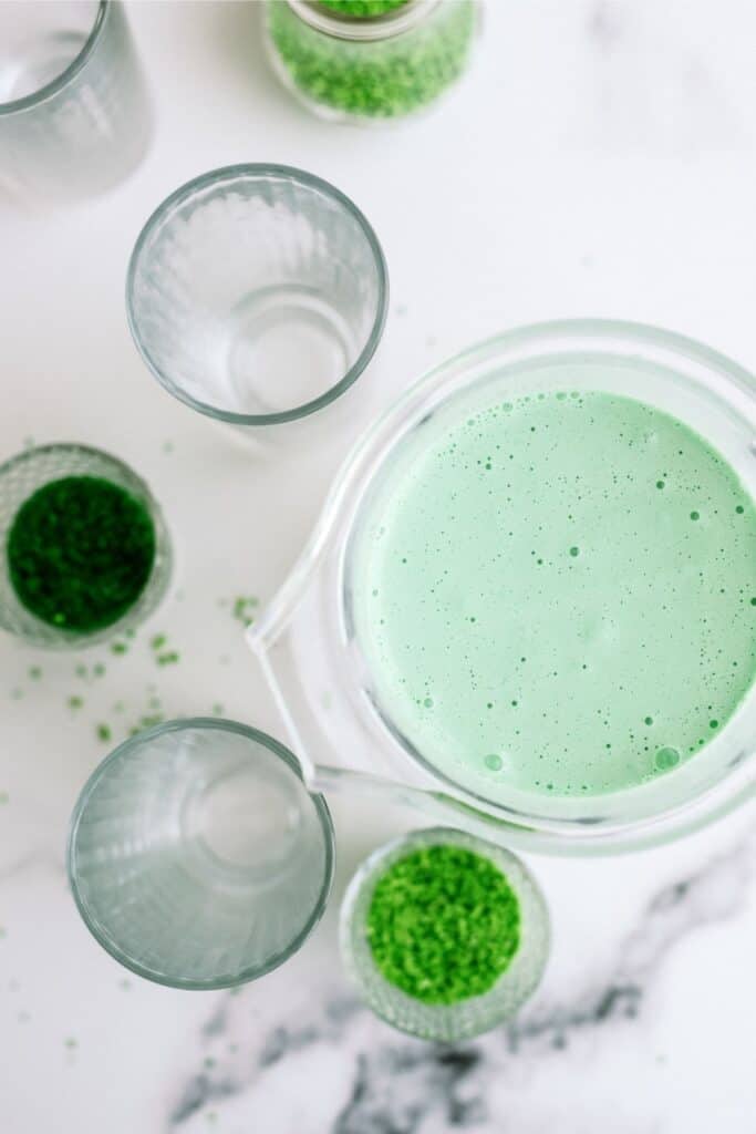 Blender with green colored ice cream next to two serving glasses and green sprinkles in a cup.