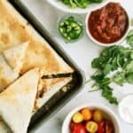 Triangular quesadillas on a baking sheet with bowls of shredded lettuce, salsa, jalapeños, cilantro, cherry tomatoes, and sour cream on a marble surface.