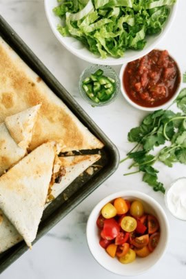 Triangular quesadillas on a baking sheet with bowls of shredded lettuce, salsa, jalapeños, cilantro, cherry tomatoes, and sour cream on a marble surface.