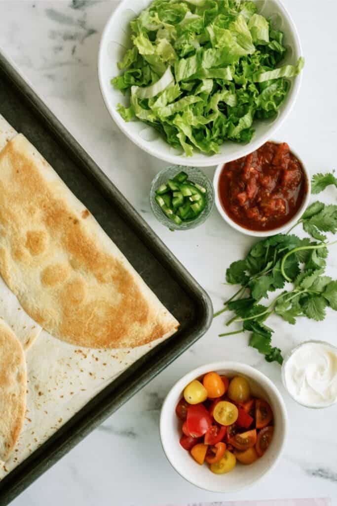 A tray with quesadillas beside bowls of shredded lettuce, salsa, cherry tomatoes, chopped jalapeños, and sour cream, with fresh cilantro on the side.