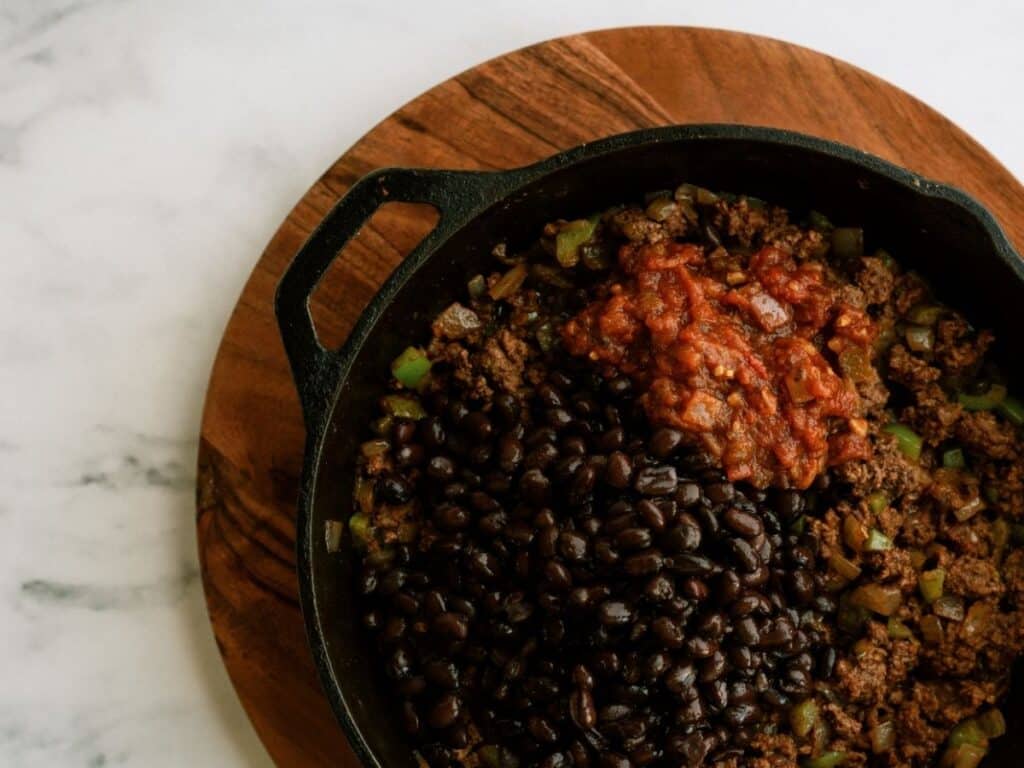 Cast iron skillet with cooked ground beef, black beans, diced green peppers, and salsa on a wooden board.