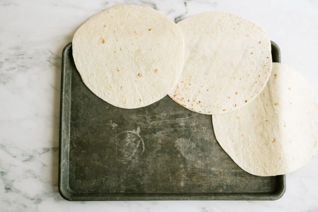 Three uncooked tortillas placed on a metal baking sheet on a marble surface.