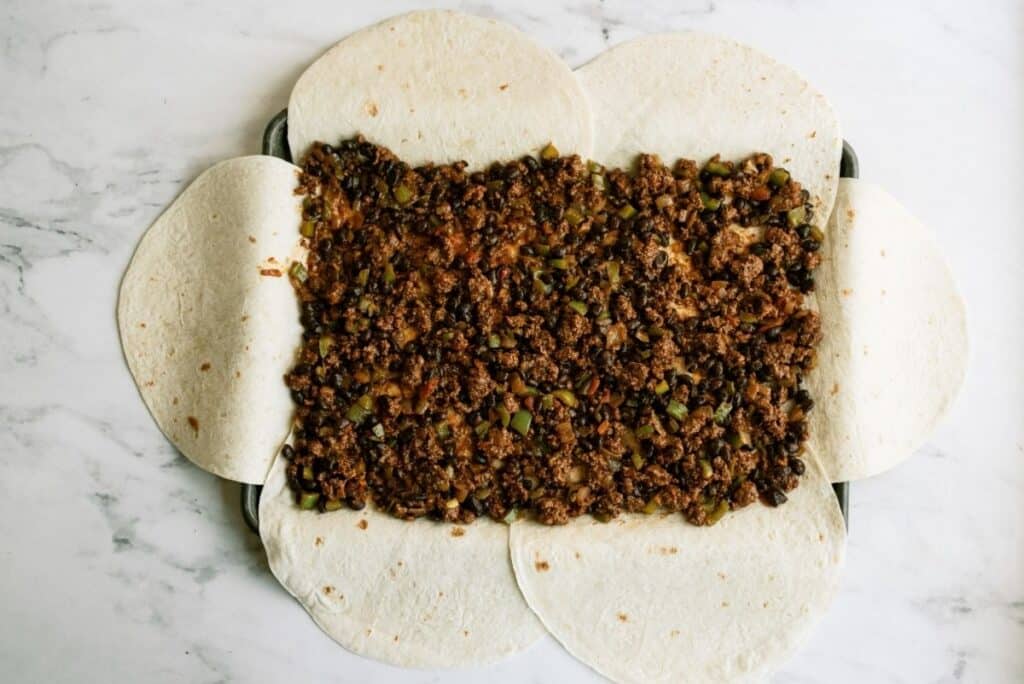 Tortillas arranged overlapping with a filling of ground meat and vegetables in the center on a marble surface.