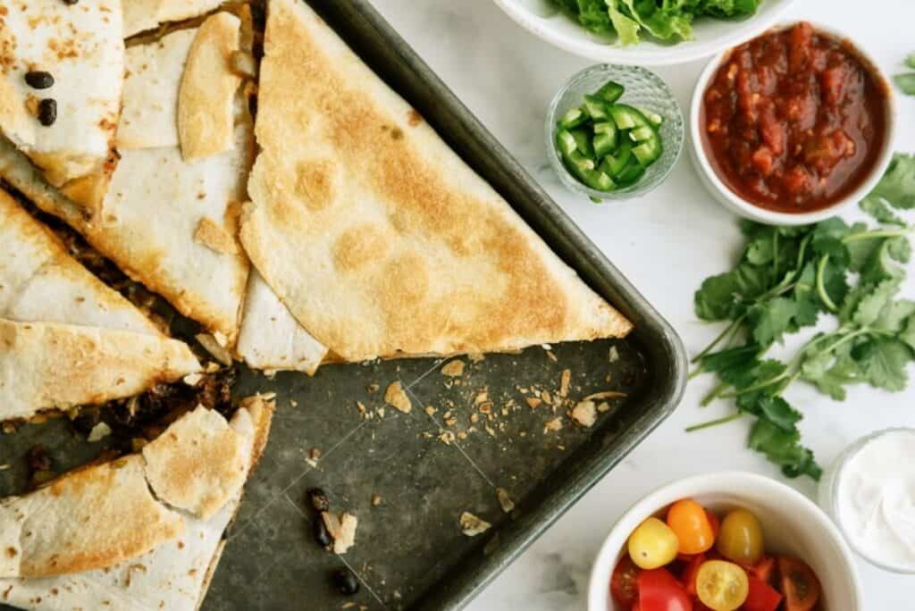 A sheet pan with sliced quesadilla, surrounded by bowls of salsa, jalapeños, lettuce, tomatoes, and cilantro on a marble surface.