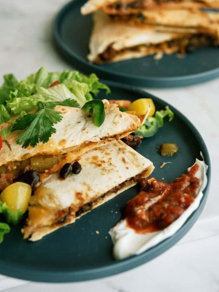 Two quesadilla slices with black beans and vegetables on a dark plate, served with a side of lettuce, sour cream, and salsa.