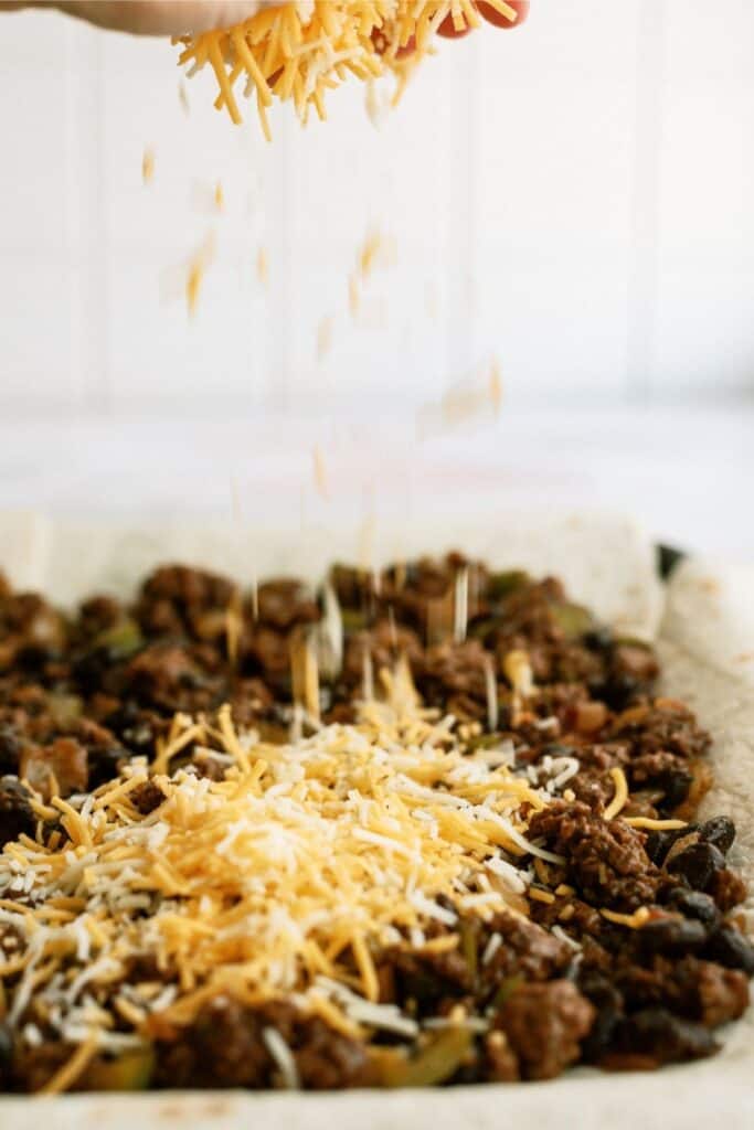 Cheese being sprinkled over a dish topped with cooked meat and vegetables on a baking sheet.