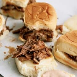 A group of pulled pork sliders on white buns with a knife placed on the white surface next to them. One slider is open, showing the pulled pork and sauce inside.