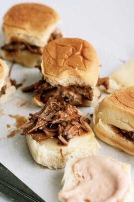 A group of pulled pork sliders on white buns with a knife placed on the white surface next to them. One slider is open, showing the pulled pork and sauce inside.