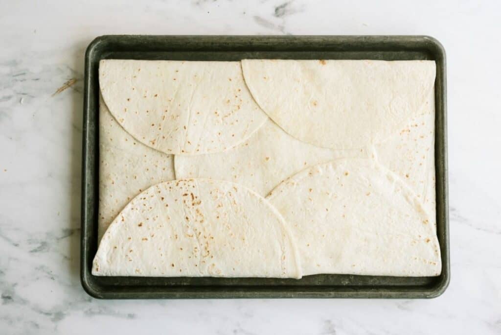 Flour tortillas folded in half arranged on a baking tray atop a marble surface.