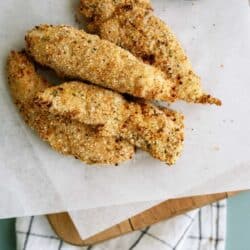 Four breaded chicken tenders on parchment paper with a cup of dipping sauce in the corner, placed on a wooden board with a cloth underneath.