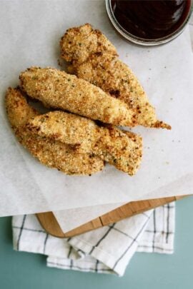 Four breaded chicken tenders on parchment paper with a cup of dipping sauce in the corner, placed on a wooden board with a cloth underneath.