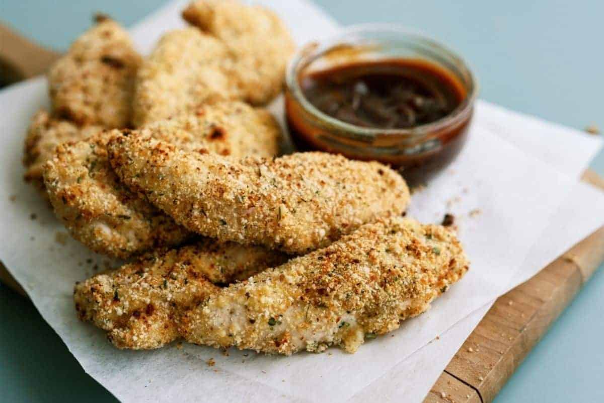 Breaded chicken tenders on parchment paper with a small bowl of sauce on a wooden board.