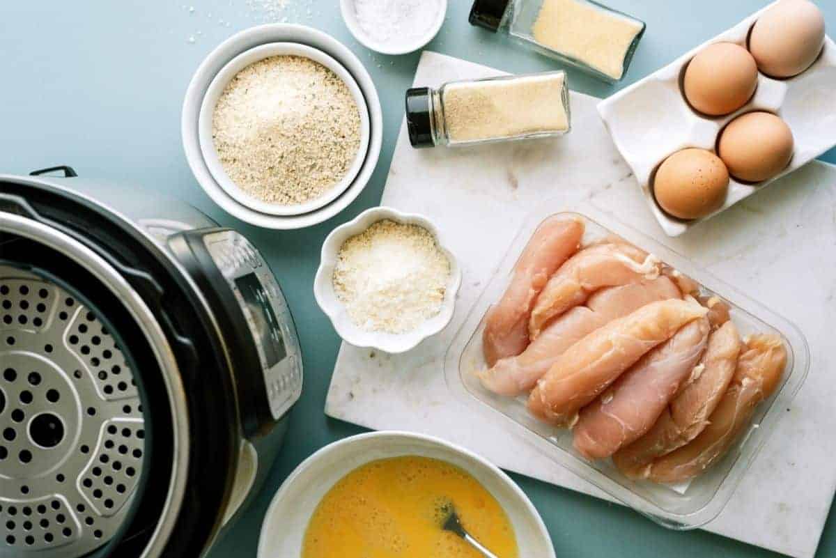 Ingredients for a breaded chicken recipe are arranged on a table including chicken breasts, eggs, breadcrumbs, grated cheese, and seasonings next to a pressure cooker.