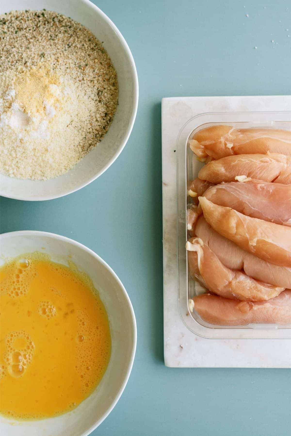 Bowls of breadcrumbs and beaten eggs beside a tray of raw chicken tenders on a blue surface.