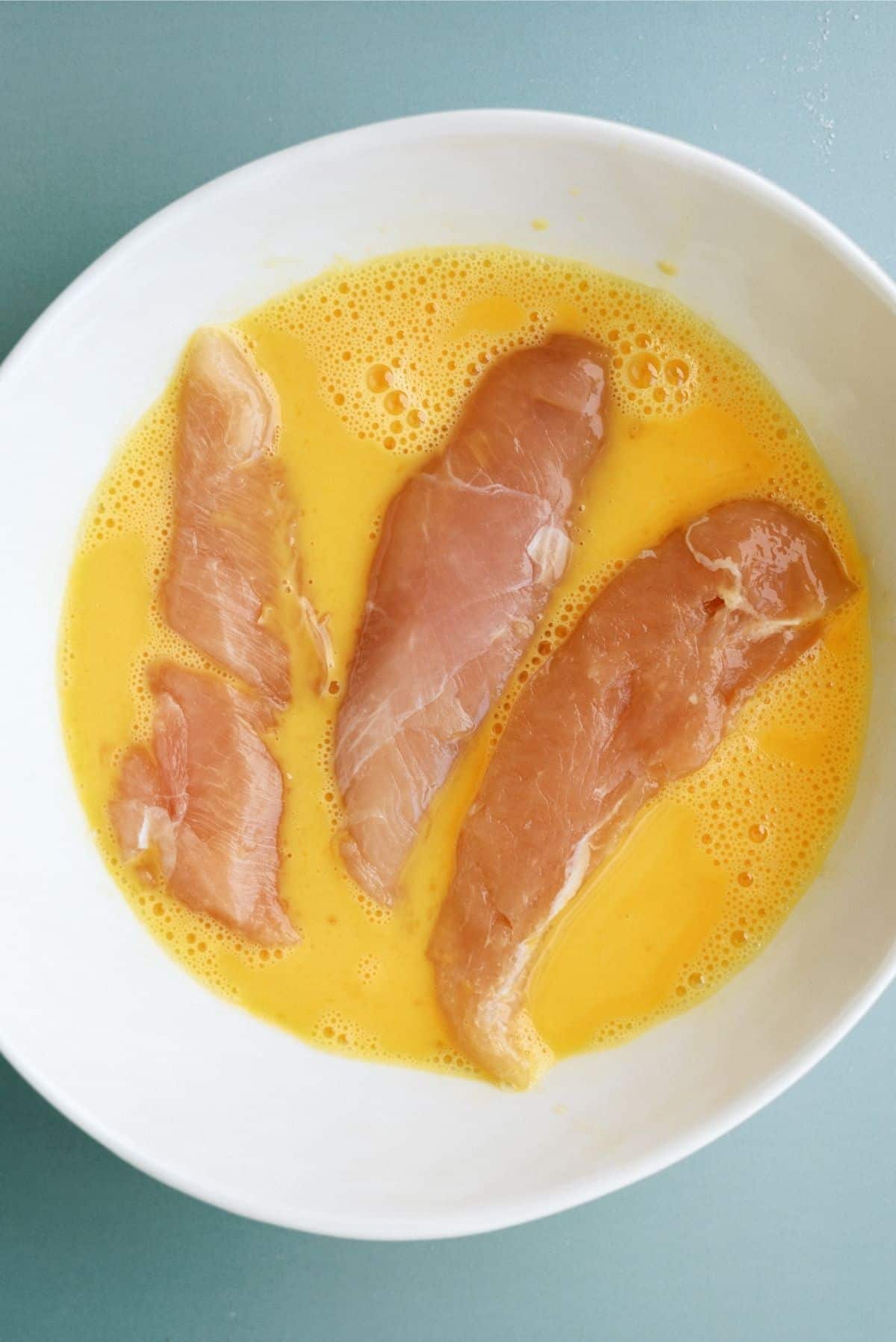 Raw chicken breasts marinating in a bowl of beaten eggs.