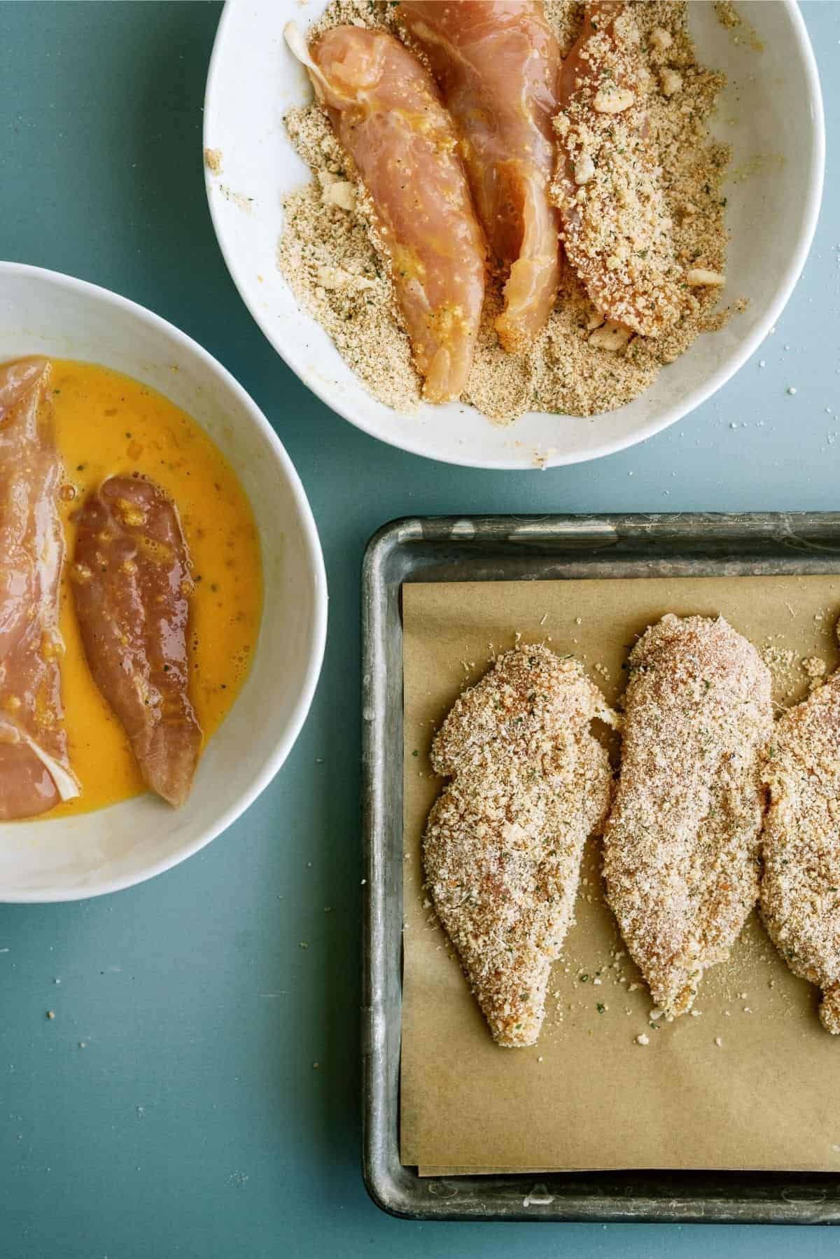 Bowls with raw chicken tenders are being coated in egg and breadcrumbs on a green surface, with breaded tenders arranged on a baking tray lined with parchment paper.