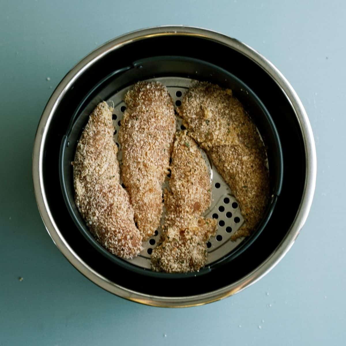 Breaded chicken strips in an air fryer basket on a blue surface.