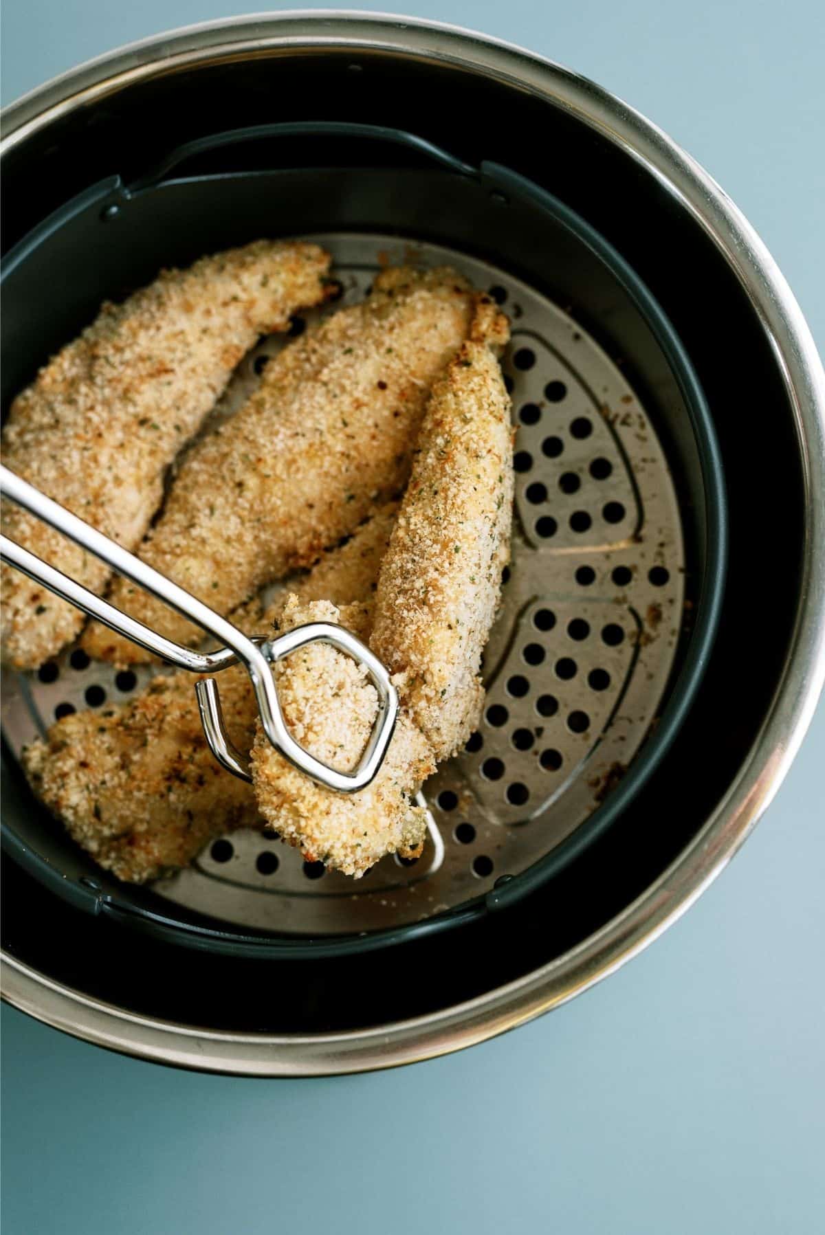 Breaded chicken tenders in an air fryer basket with metal tongs gripping one piece.