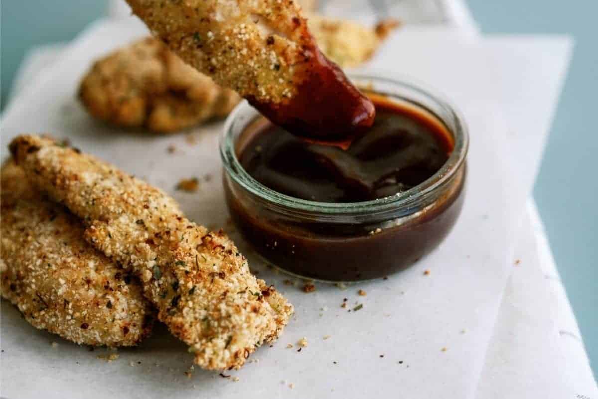 Breaded chicken is being dipped into a small glass bowl of barbecue sauce. Other pieces are on a paper-lined surface.