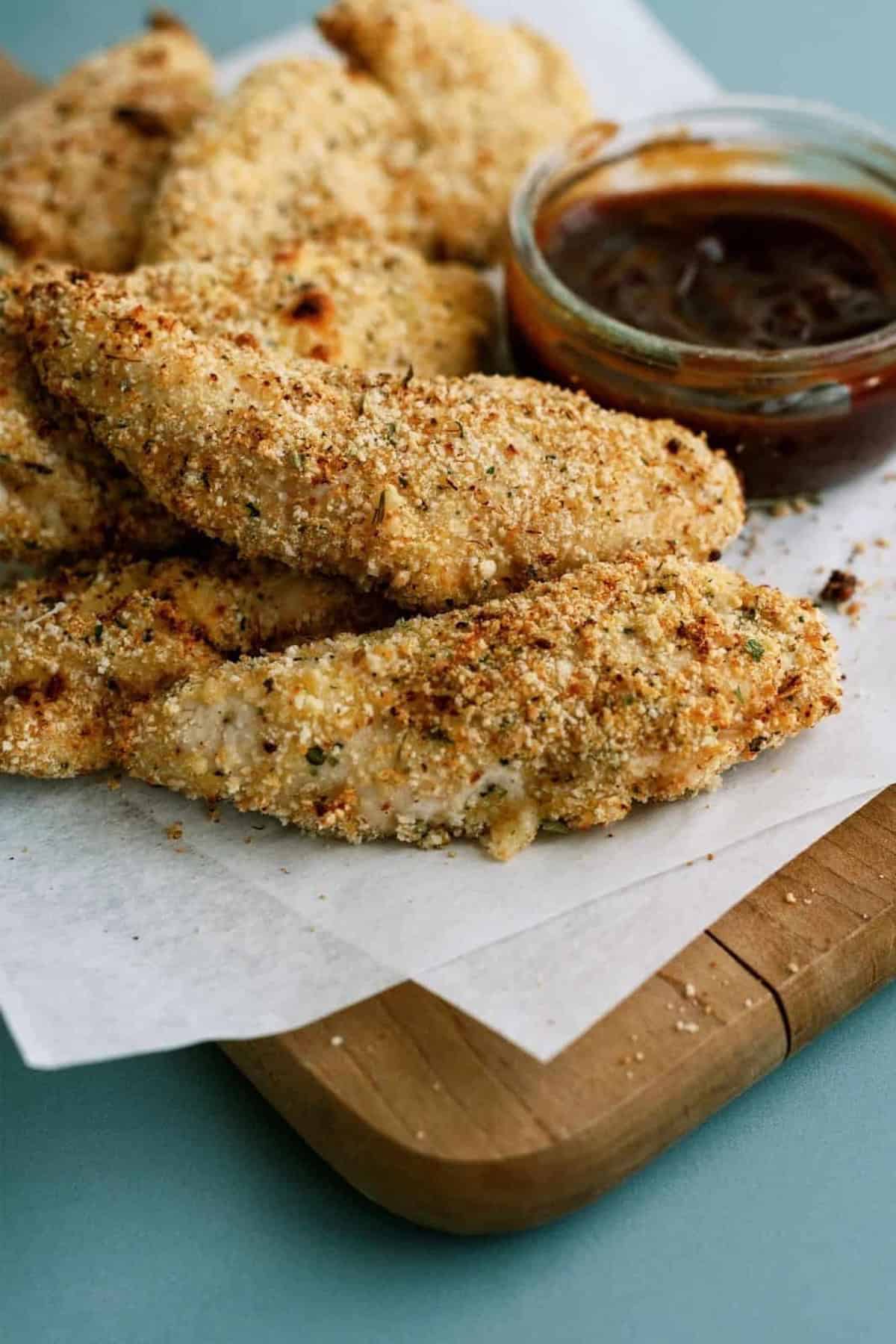 Breaded chicken tenders on parchment paper with a side of barbecue sauce on a wooden cutting board.