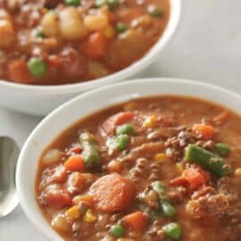 Two bowls of hearty beef and vegetable soup with visible pieces of carrots, potatoes, green beans, peas, and corn. A silver spoon is placed beside the bowls on a light surface.